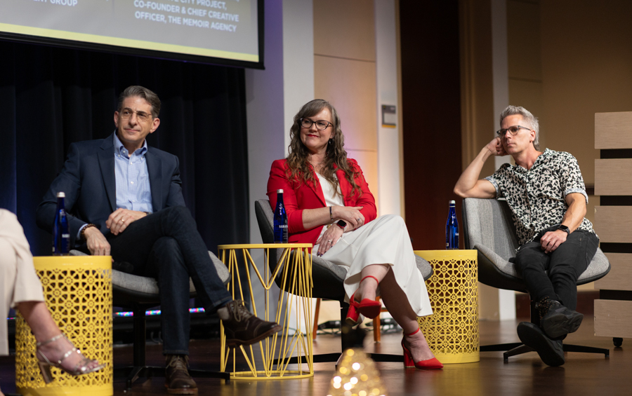 Three panelists seated on stage in armchairs: a man in a navy suit, a woman in a red blazer and white dress, and a man in a patterned shirt, with blue water bottles on nearby yellow tables.