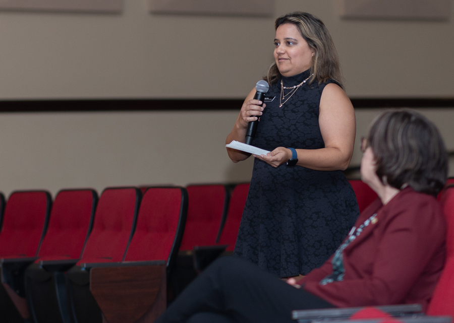 Woman in a dark lace dress speaks into a handheld microphone, holding notes in a conference setting with red seats behind her.