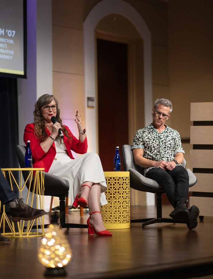 Woman in red blazer speaks into a microphone on a panel stage, seated beside a man in a patterned shirt; a yellow lattice table and water bottles nearby.