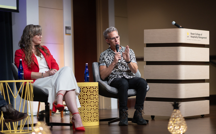 Panel discussion: woman in red blazer and white dress and man in patterned shirt speak on stage with a microphone, podium on the right labeled Rosen College of Hospitality Management.