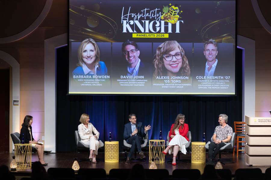 Five panelists seated on a stage during a conference, with a large screen behind them reading 'Hospitality Knight Panelists 2026'. The lineup includes a woman in black, a woman in a white suit, a man in blue, a woman in red, and another woman in a patterned top, with small gold tables in front of them and blue stage lighting.