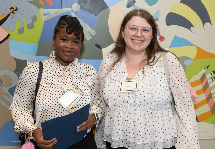Two women smile for a photo, standing side by side in front of a bright abstract mural; the woman on the left holds a blue folder and wears a polka-dot blouse with a name badge.