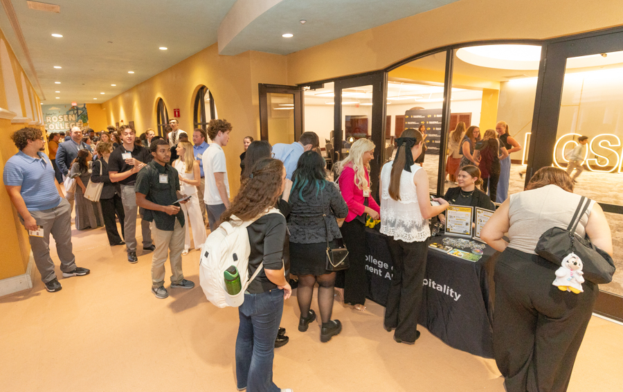 Crowded campus hallway with attendees lined up at an information booth labeled Hospitality, gathering brochures and talking to staff.