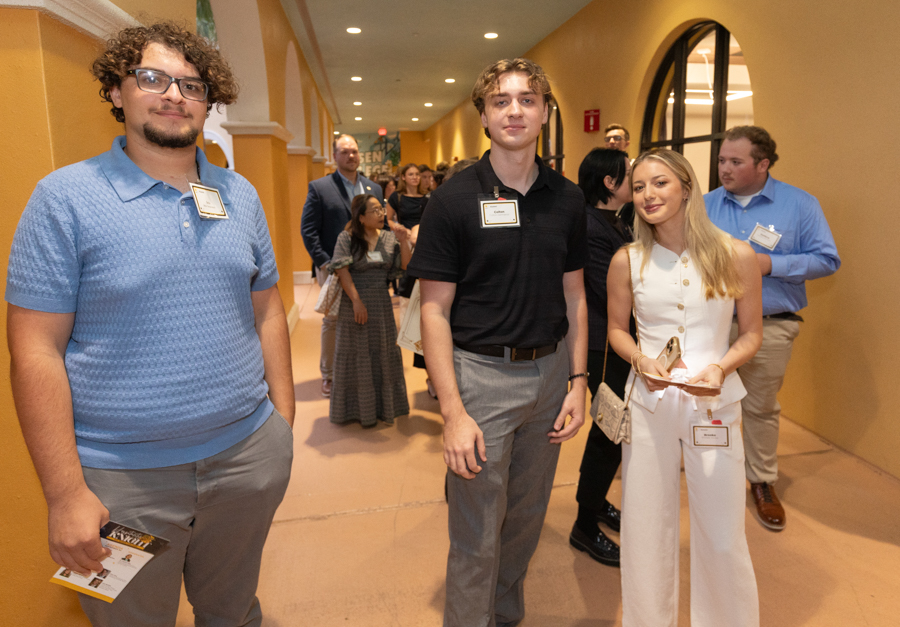 Three young adults with name badges pose in a conference hallway; left man in a blue polo, center man in black shirt, right woman in white suit.