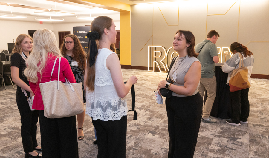 Group of people chatting in an indoor event space, with a warm-toned wall and large lit decorative letters in the background.