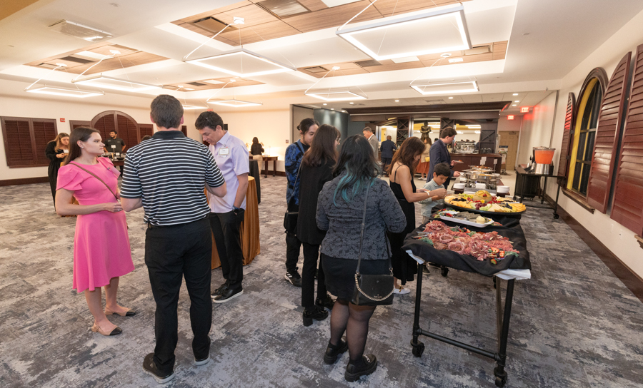 Group of people mingling at a buffet in a bright banquet hall, with tables of appetizers on the right.