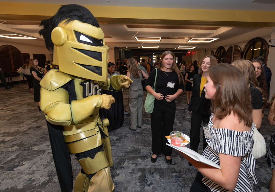 Golden knight mascot greets a student group at a campus event; a girl in a striped top holds a plate with a drink while others chat nearby.