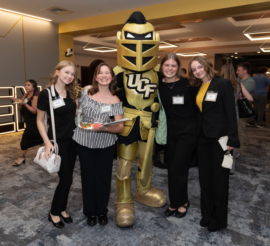 Four women in semi-formal attire pose with a gold armored knight mascot bearing 'UCF' at a social event in a modern lobby, some holding plates of food and badges visible.
