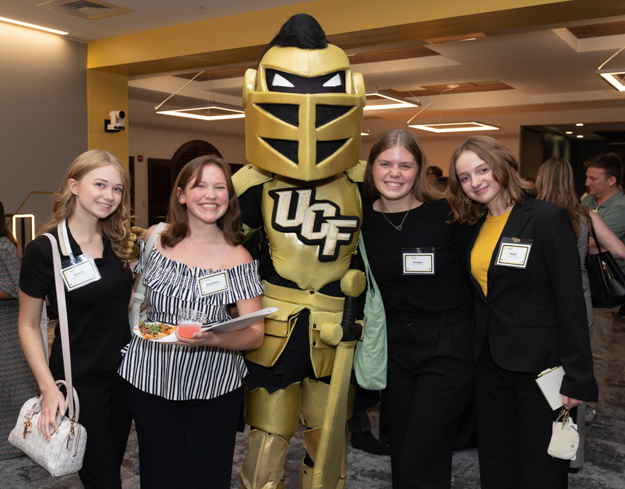 Four young women posing with a gold UCf Knights mascot at a social event. They smile and wear name tags in a well-lit indoor venue.