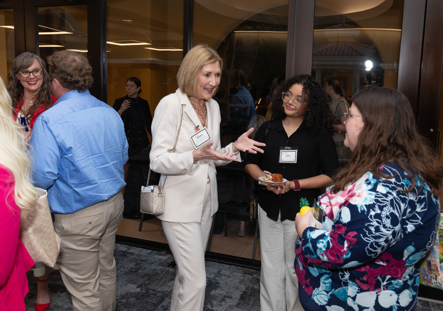 Business networking event: woman in white suit speaks with hand gestures while others listen, name tags visible.