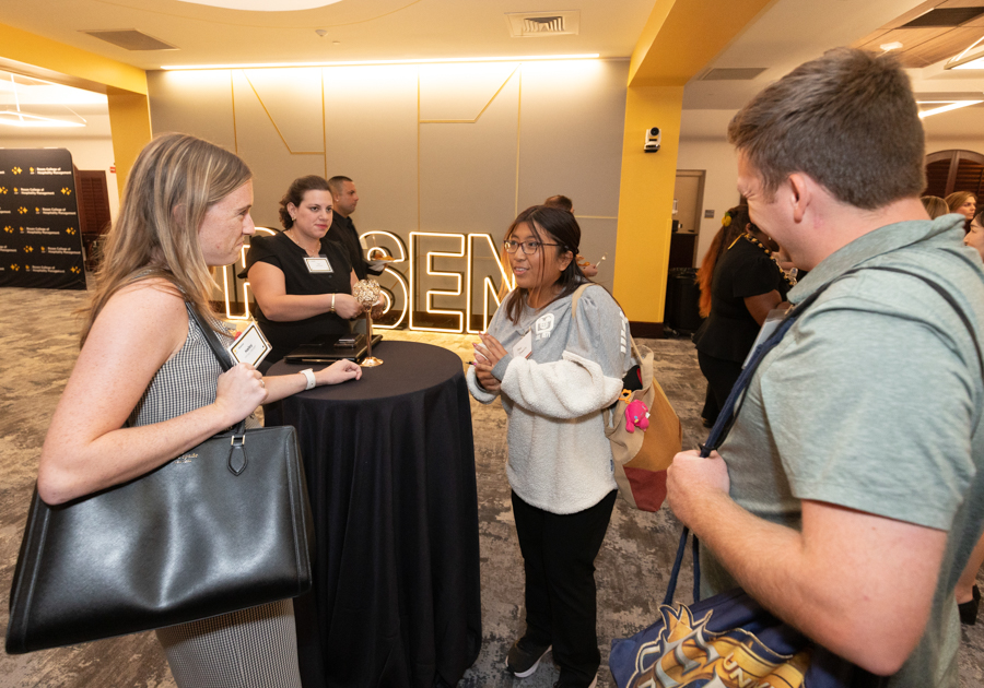 Three attendees and an staffer chat around a tall round table at a conference reception, with large illuminated letters in the background.