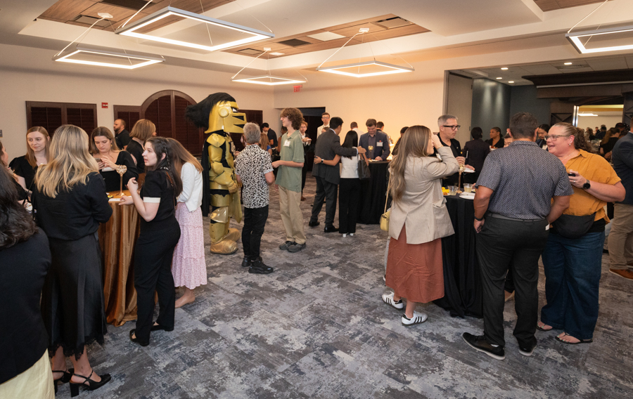 Guests mingle at a reception in a conference venue, with a person in a shiny gold mascot costume at the center.