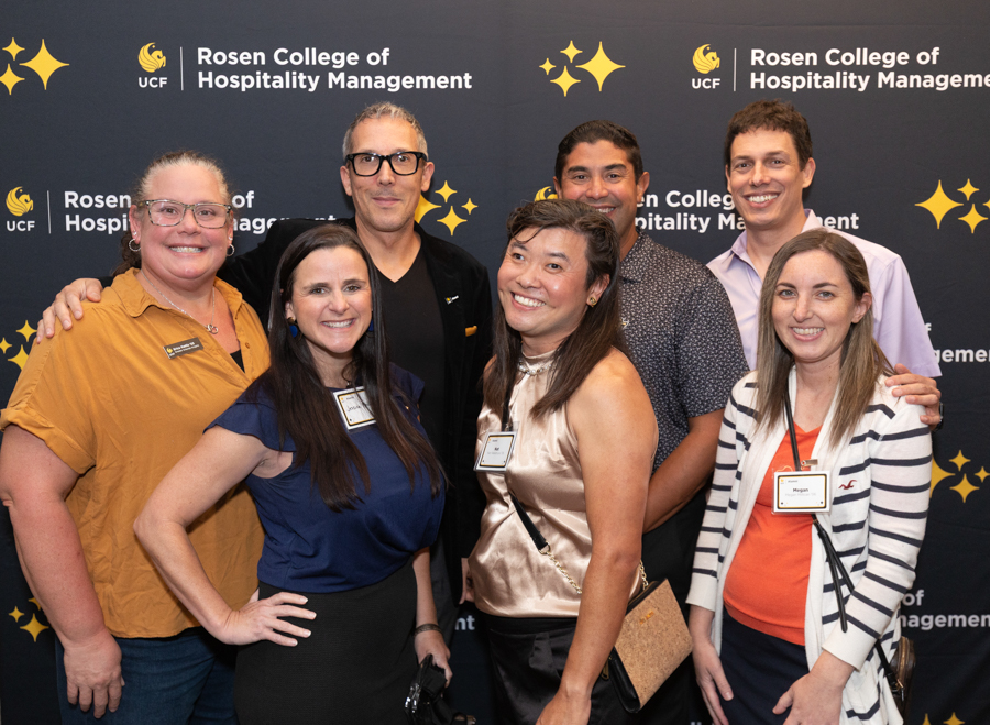 Group of six smiling professionals posing for a photo at a Rosen College of Hospitality Management event backdrop.