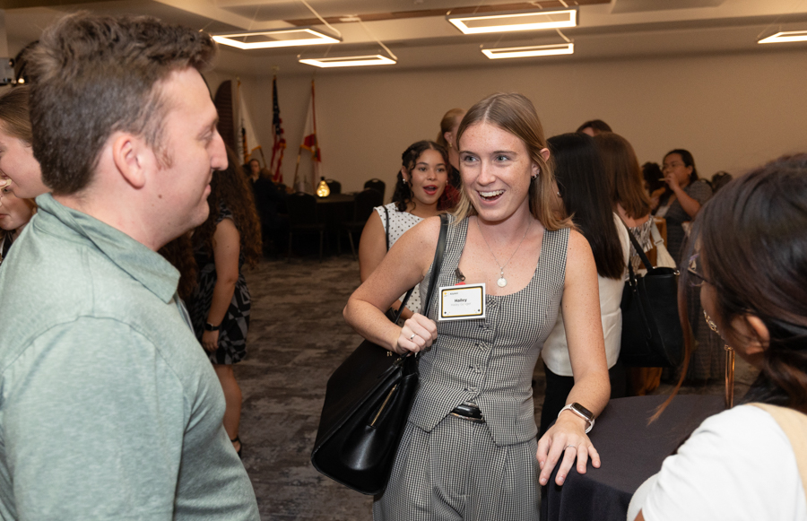 Smiling woman in a checkered dress at a networking event talks to a man, name badge visible on her chest.