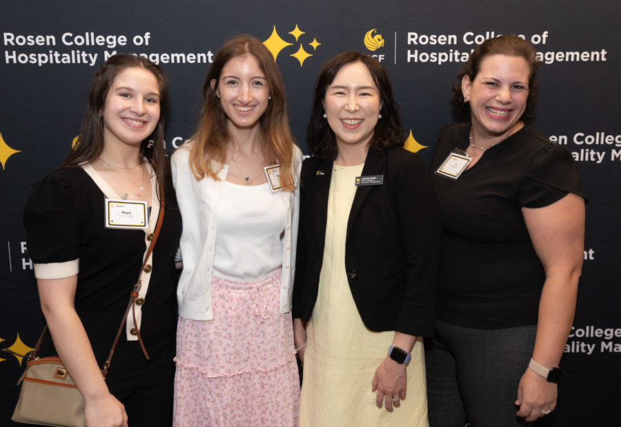 Four women posing together at a Rosen College hospitality event, smiling in front of a backdrop with the college logos and branding.