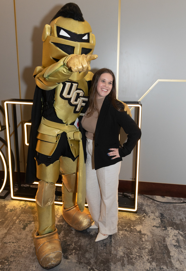 Woman smiles while posing with a tall gold knight mascot that points toward the camera at an indoor event.