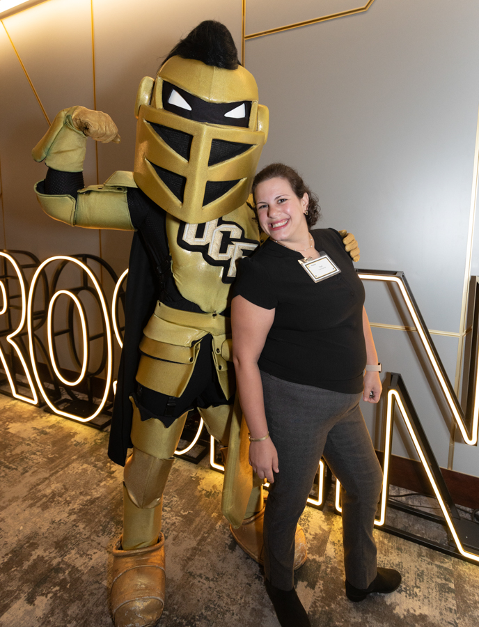 Smiling woman posing with a large gold knight mascot in armor indoors.