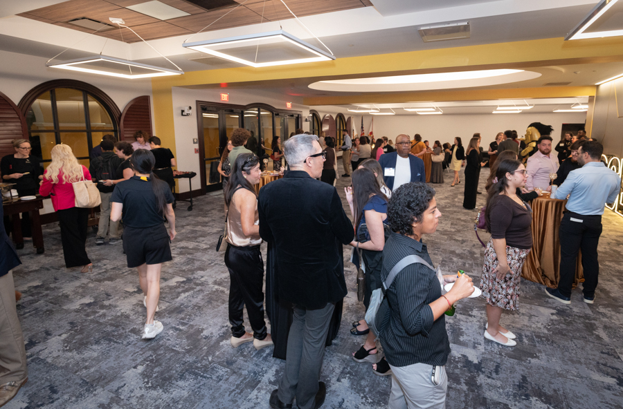 People mingle at a reception in a large, well-lit event hall with gray carpeting and orange accents, round tables, and a diverse crowd of guests in smart-casual attire.