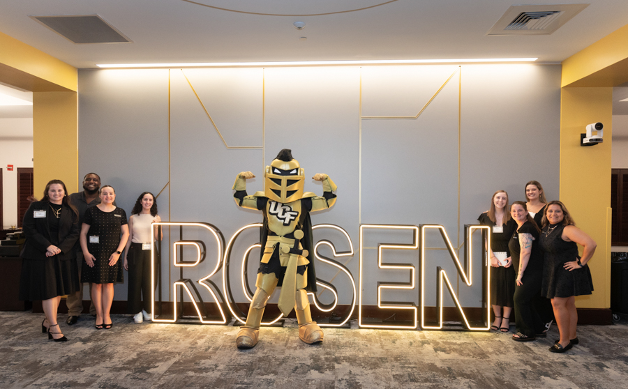 Group of people posing with a gold mascot in front of a large illuminated sign that reads 'ROSEN'.