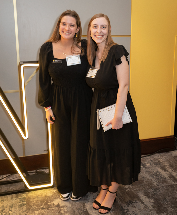 Two women in black dresses pose together at a formal event, smiling; one holds a white studded clutch and name badges are visible.