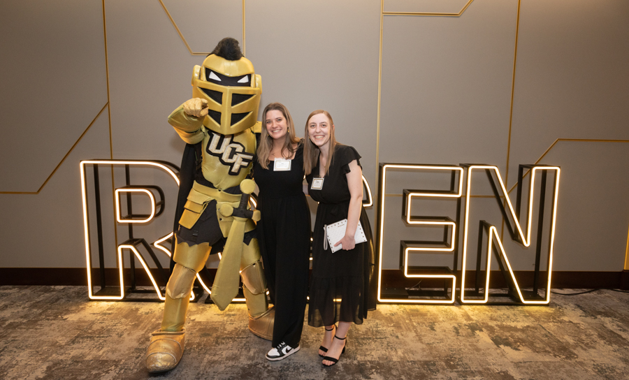 Two women pose with a gold knight mascot at a conference, standing in front of a glowing sign.