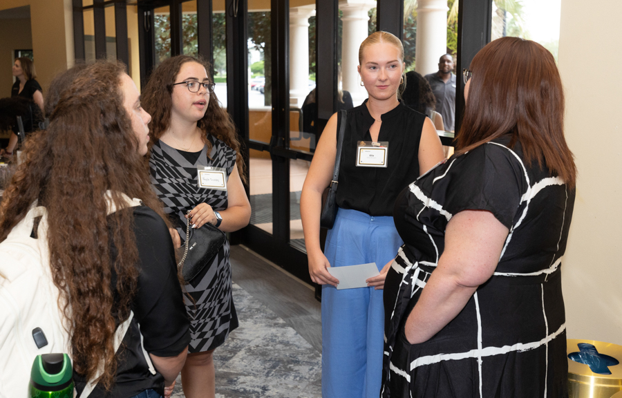 Four women converse in a hallway at an event, wearing name badges on their chests.