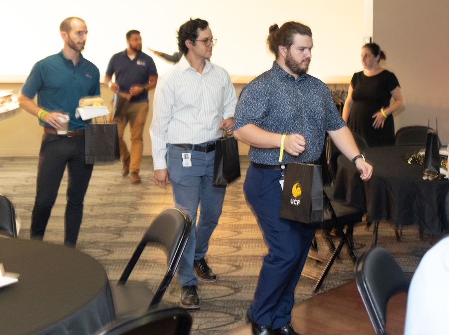 Group of professionally dressed attendees carrying gift bags walk through a conference room, with tables set for an event.