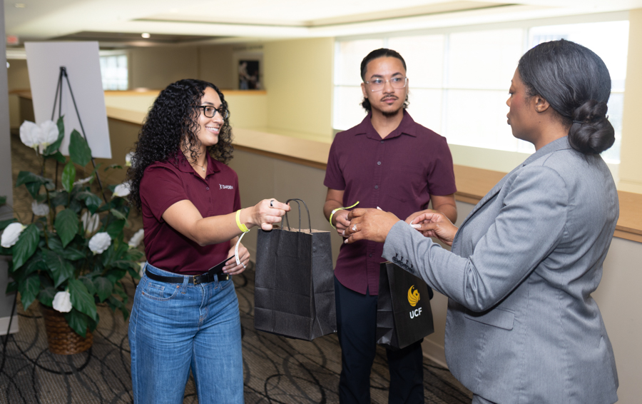 Staff member in a maroon shirt hands a black gift bag to a guest in a gray suit at a campus reception area, with a man observing nearby.