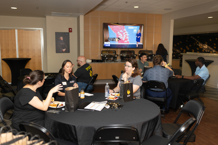 People sit at round tables with black tablecloths, eating and talking in a lobby as a large monitor shows a map presentation on the wall.