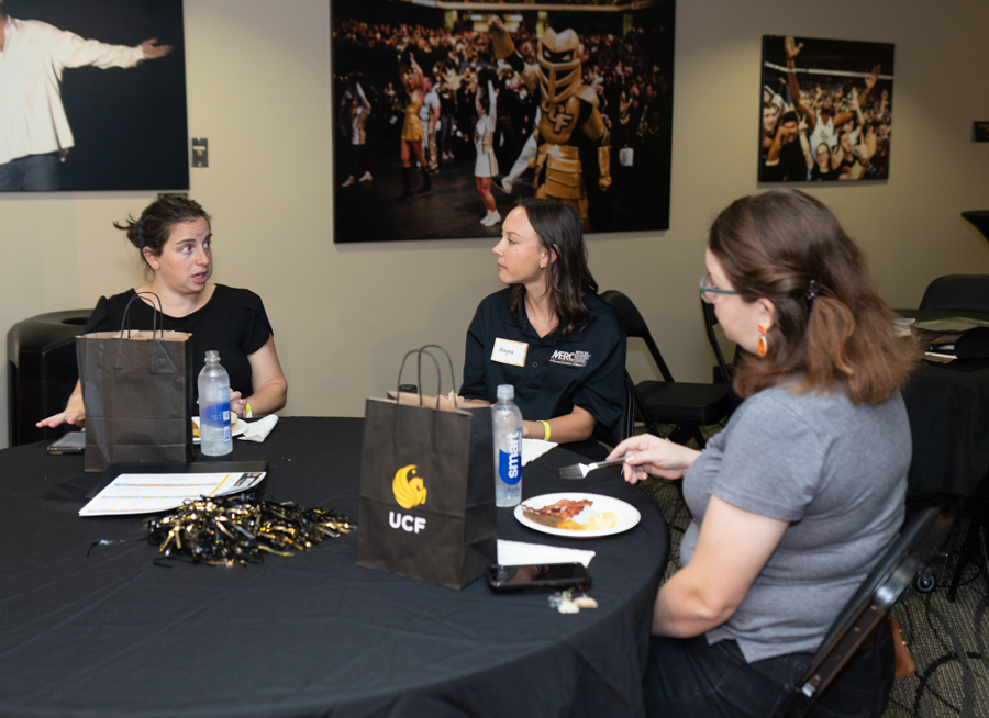 Three women sit around a black-clothed table at a campus event, with a UCF-branded bag, water bottles, and snacks visible.