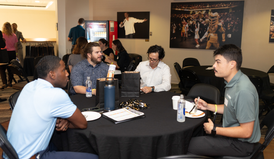 Four men sit around a round table at a casual event, eating and chatting with a gift bag and beverages on the table.