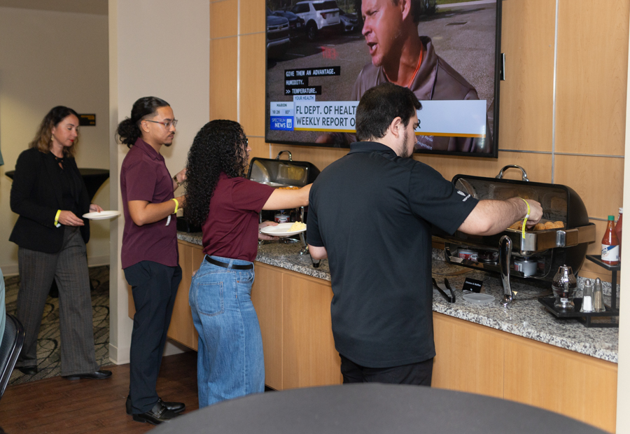 People line up at a buffet station with chafing dishes, serving themselves; a TV screen is mounted on the wall above the counter.
