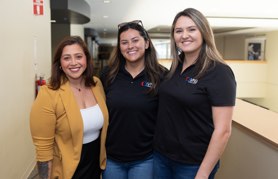 Three smiling women pose together in a bright office hallway, two in matching black logo polo shirts and one in a mustard blazer.