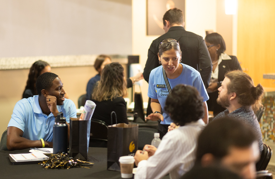 Facilitator in a blue shirt chats with attendees at a table during a community event, with gift bags and cups nearby.