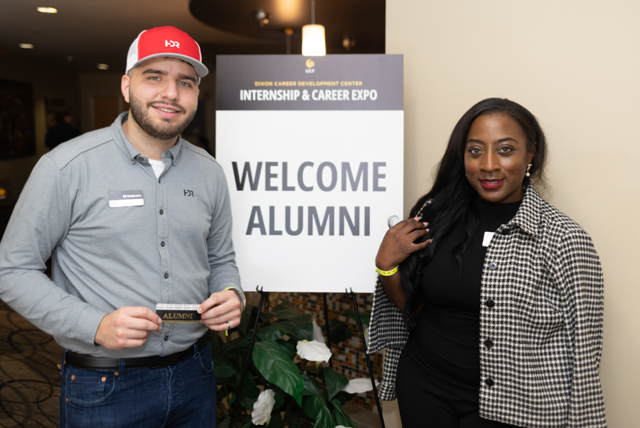 Two attendees stand beside a sign reading 'Welcome Alumni' at an internship and career expo, one man holding an alumni badge.
