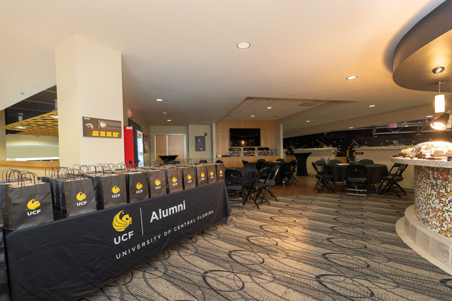 Black gift bags with UCF Alumni logo lined up on a table in a modern lounge; seating and a curved bar in the background.
