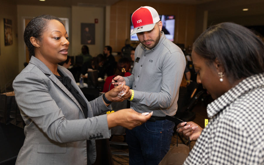 Staff member in a gray blazer hands a badge to a guest at a busy event check-in. A man in a red cap and others work in the background on wristbands.