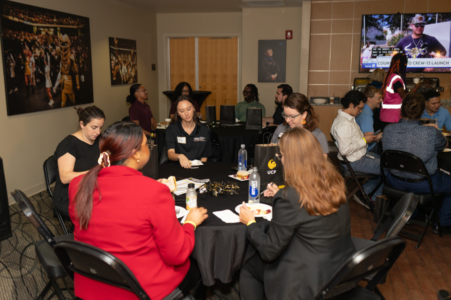 Diverse adults share a meal at a round table in a conference room during a social event, with more attendees and a TV screen in the background.