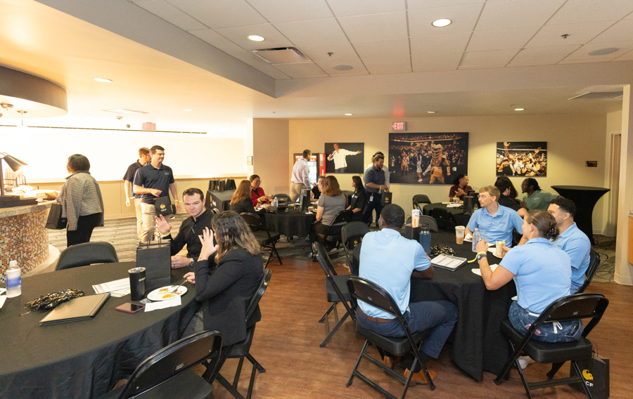 Group of people seated at round tables with black tablecloths in a casual corporate reception area, chatting and eating snacks.