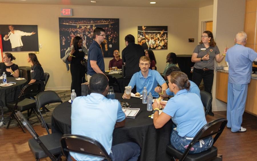 People seated around round tables eating and chatting at a casual networking event in a modern conference room with wall art and an exit sign visible in the background.