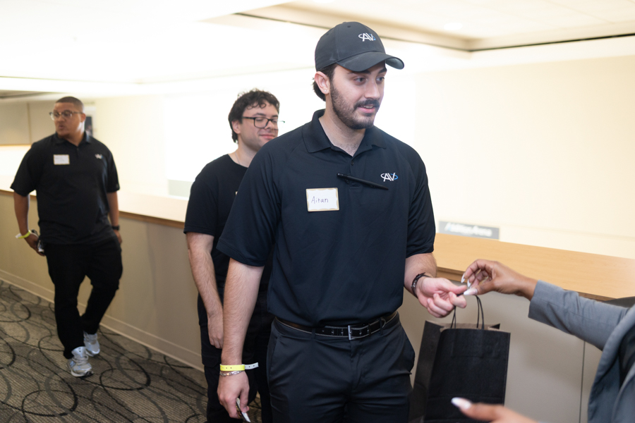 Man in a navy polo with a name tag labeled 'Aitan' hands a small item to someone off-screen while two teammates walk behind indoors in a hallway/reception area.
