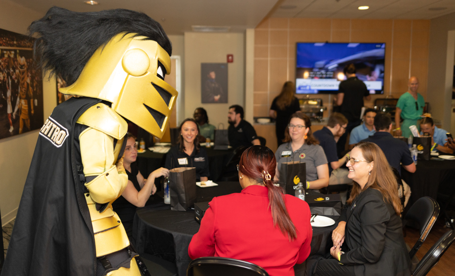 Gold knight mascot converses with professionals at round tables during a conference reception.