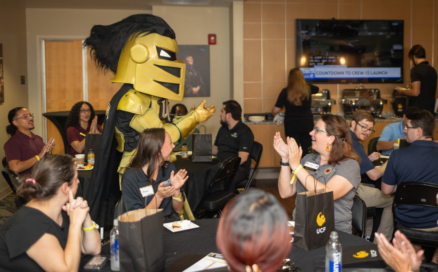 Gold knight mascot walks between tables as attendees clap at a university event.
