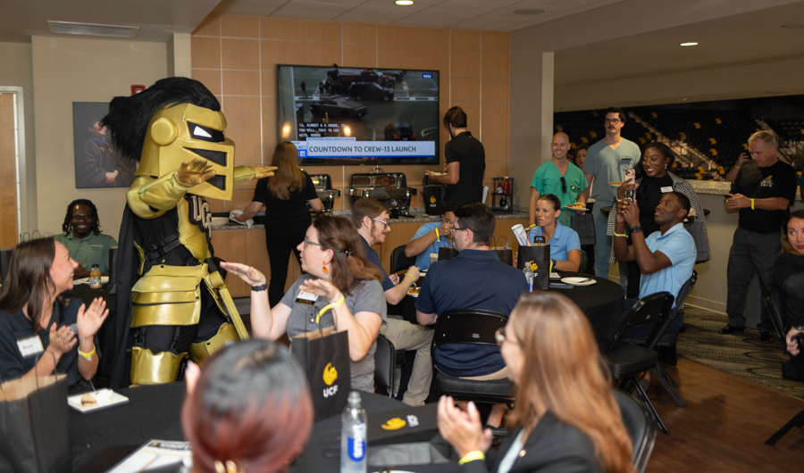 Gold armored mascot greets and interacts with guests at a casual event, while attendees at round tables applaud and chat.