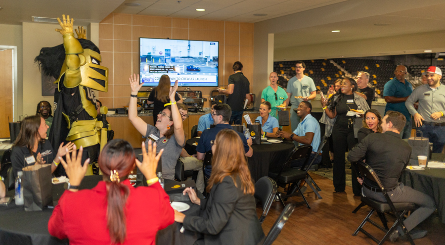 Coworkers at a reception raise hands and smile around round tables; a gold-armored mascot stands at left as a TV screens shows in the background.
