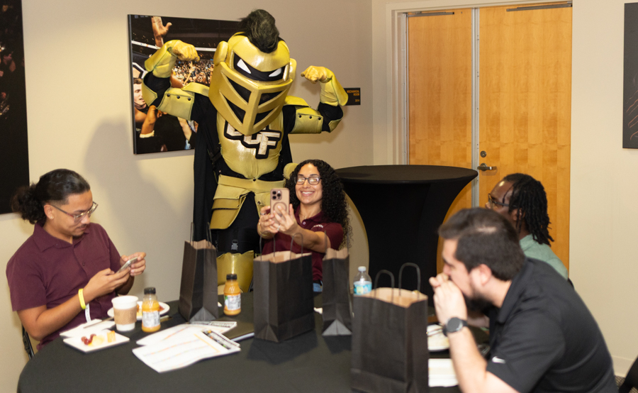 People gathered around a round table at an event, a gold armored mascot posed behind them as someone snaps a selfie with a phone.