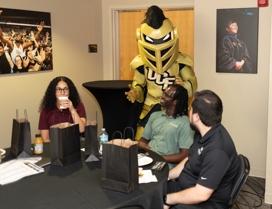 Group of four people seated around a round table with black gift bags; a gold armored mascot stands behind them, smiling and engaging with the group.