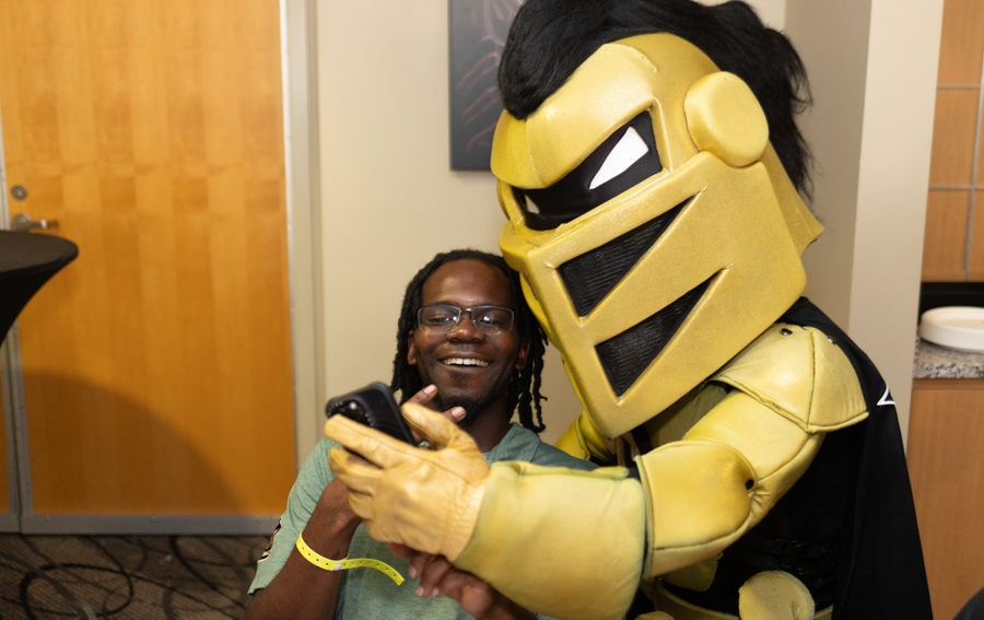 Man in a green shirt smiles for a selfie with a gold knight mascot, holding a phone together.