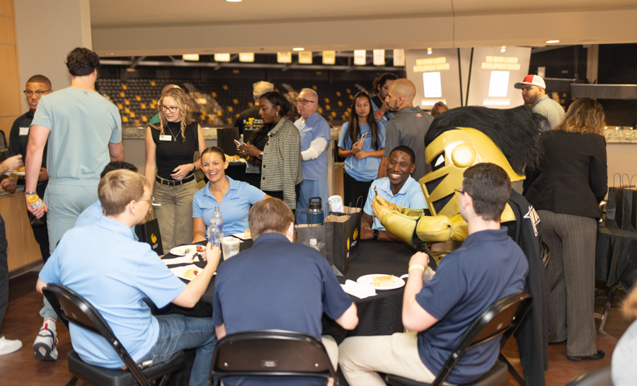 Group of people at a casual event around a round table, some in blue shirts, mascot in a gold suit greeting attendees.