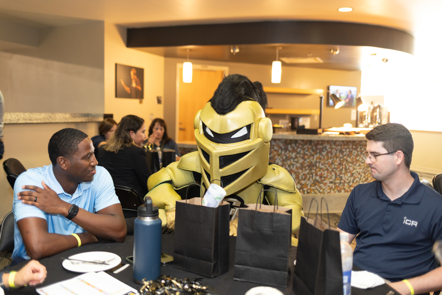 Person in a gold knight costume sits at a table with two men in a casual restaurant scene, black gift bags and plates on the table in the foreground; warm lighting in the background.
